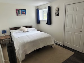 A bedroom with a bed, a nightstand, a heater, a window with curtains, and a door.