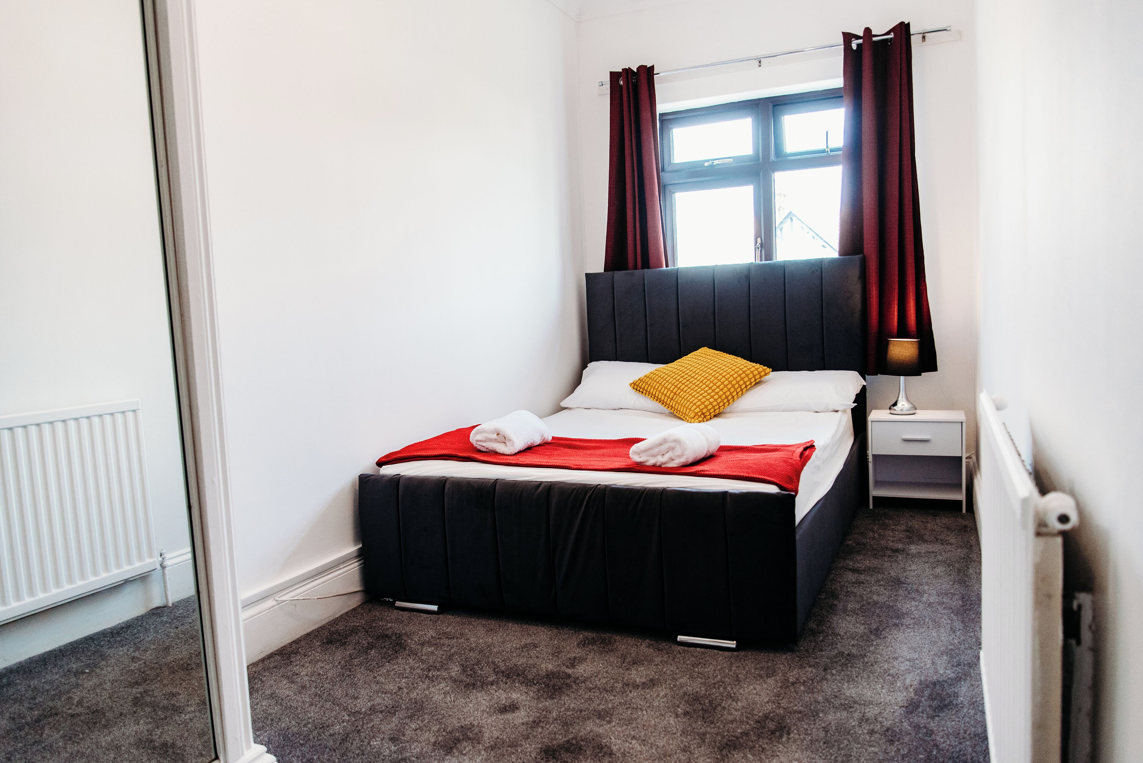 A neatly made bed with a black headboard and a yellow pillow, a white bedside table with a lamp, and a window with red curtains.
