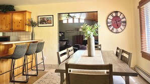 Un coin repas comprenant une table en bois, des chaises, un ventilateur de plafond, une horloge, ainsi qu’un espace cuisine avec des placards en bois et un four à micro-ondes.