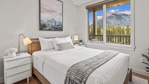 Un dormitorio con una cama grande, una mesita de noche con lámpara, un cuadro enmarcado y una ventana con vistas a la montaña.