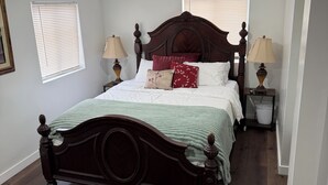 Una cama bien hecha con cabecera de madera, dos mesitas de noche con lámparas y una ventana con persianas.