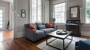 Un salon avec un canapé gris, une table basse et un fauteuil bleu. Il y a deux fenêtres avec des rideaux blancs, un miroir et une petite table avec une lampe.