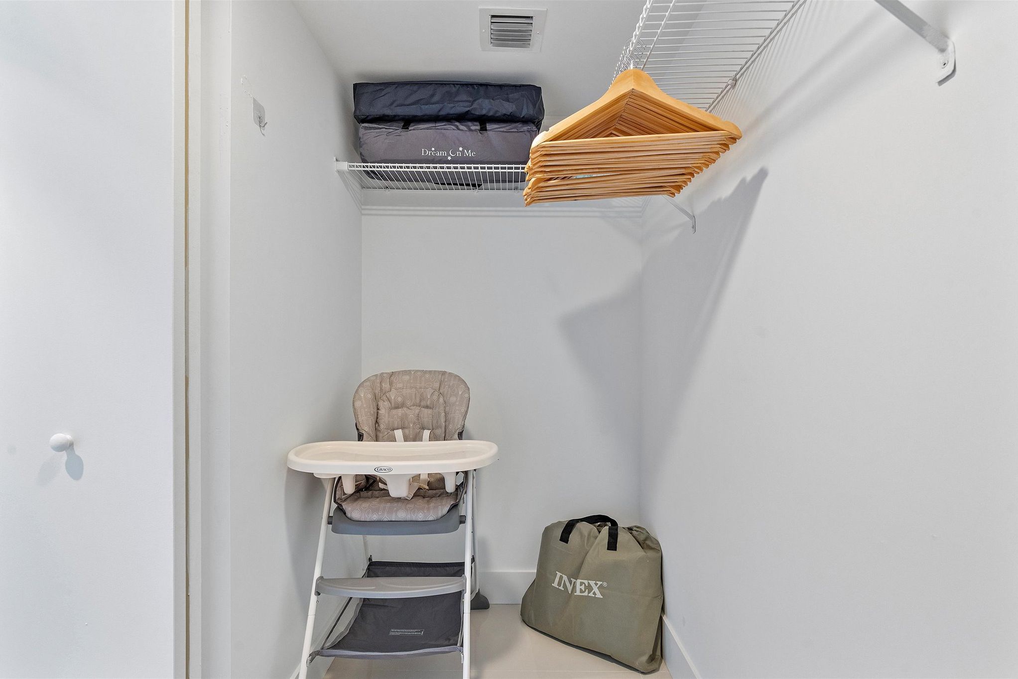 Una stanza piccola con una parete bianca, una porta bianca, uno scaffale bianco con vestiti piegati, un'alta sedia per neonati e una borsa per bambini.