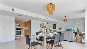 Eine moderne Küche mit Edelstahlgeräten, ein Essbereich mit Glastisch und schwarzen Stühlen sowie ein Wohnzimmer mit grauem Sofa und Fernseher.