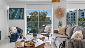 Ein modernes Wohnzimmer mit einer Couch, Sesseln, einem Couchtisch und einem großen Fenster mit Blick auf Gebäude und Bäume.
