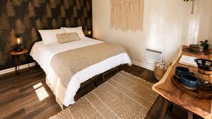 Quarto com cama, um painel de macramé, planta pendurada, mesa de cabeceira de madeira com cafeteira e tapete estampado.