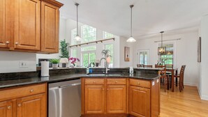 A modern kitchen with wooden cabinets, stainless steel appliances, and a large island. There are pendant lights and a dining area with wooden chairs and a table. - Hera Haven: Pickleball, hot tub, racetrack & lake (Stillwater)