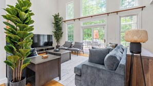 A spacious living room with a grey sofa, a coffee table, and a TV. There are large windows and a balcony visible. - Hera Haven: Pickleball, hot tub, racetrack & lake (Stillwater)