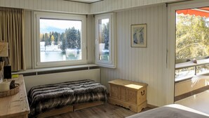 A bedroom with a bed, a wooden chest, a window with a view of a lake and trees, and a picture on the wall.