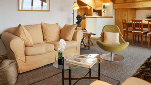 Een gezellige woonkamer met een beige bank, een glazen salontafel en een groene fauteuil. Een ingelijst beeld hangt aan de muur, en op de tafel staat een vaas met bloemen.