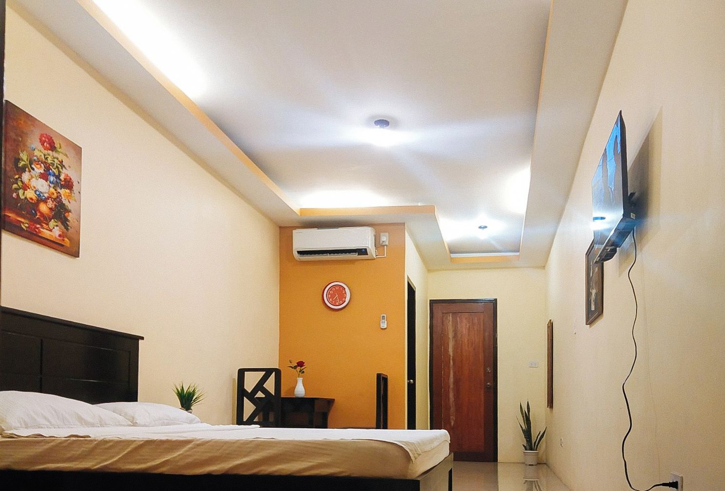 A hotel room with a bed, a desk, a chair, a clock, an air conditioning unit, a TV, and a door.