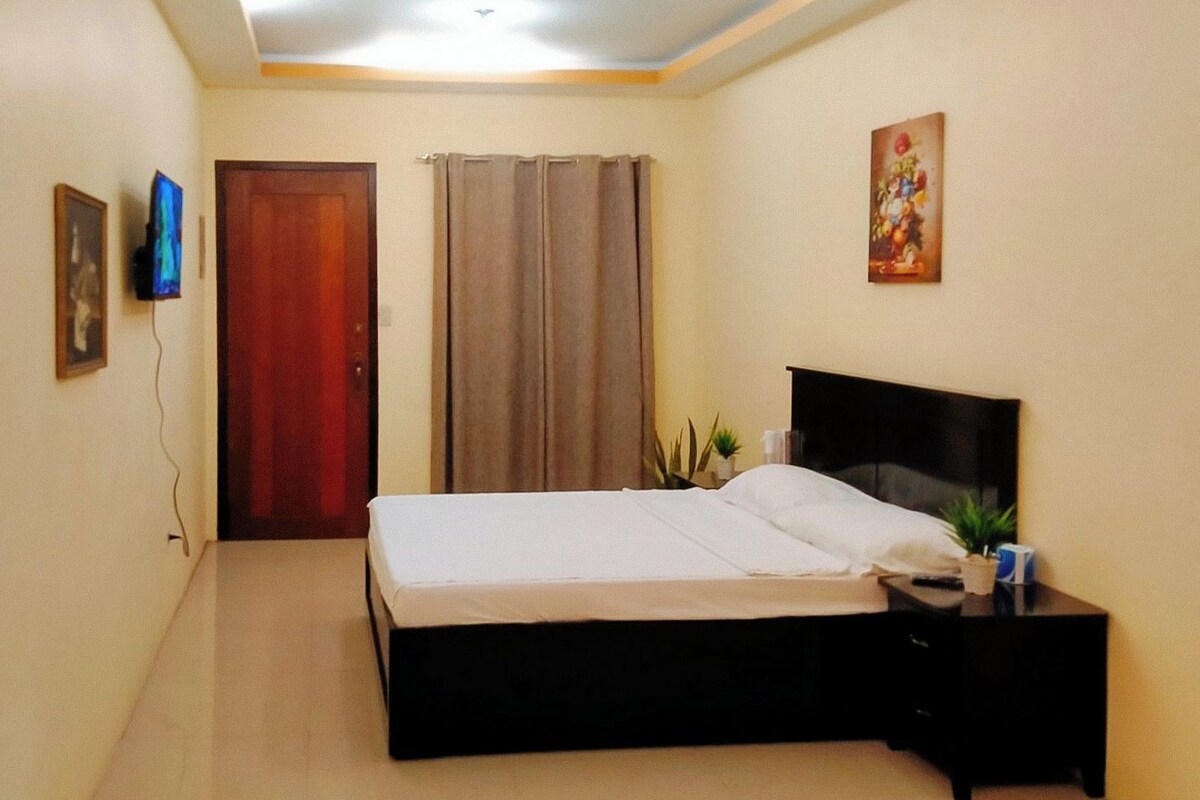 A bedroom with a bed, a nightstand, a TV, a door, and a painting on the wall.