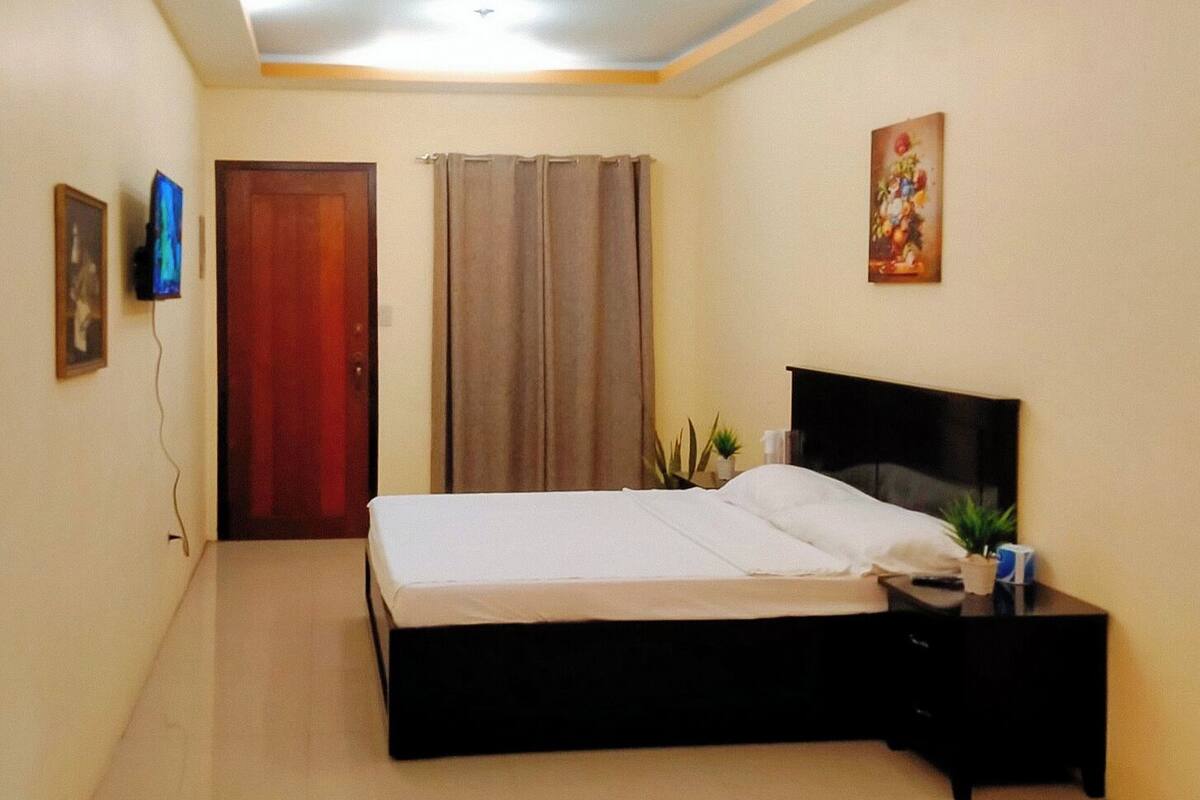 A bedroom with a bed, a nightstand, a TV, a door, and a painting on the wall.