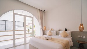 A modern hotel room with a large bed, a round wooden side table, and a view of the outdoors through a large window. - Casa Nata Graceful 2 BR Mediteranian (Canggu)