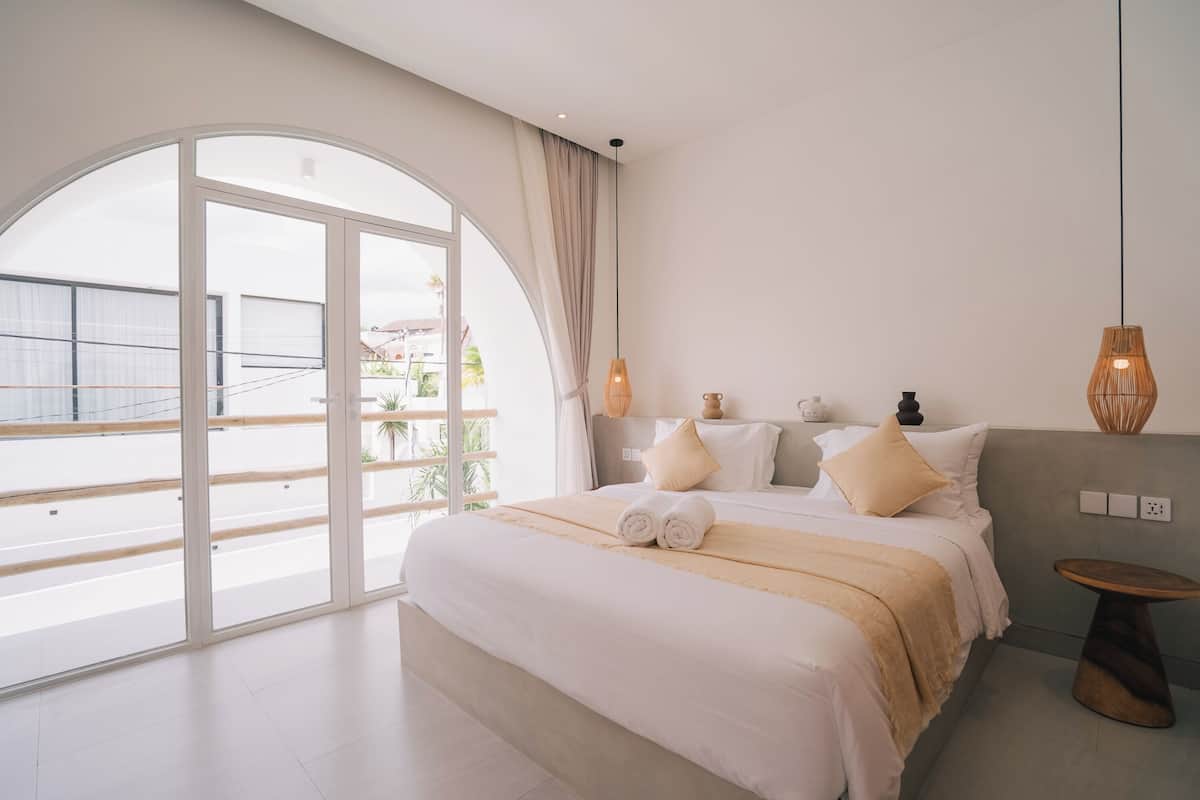 Una habitación de hotel moderna con una cama grande, una mesita redonda de madera y vistas al exterior a través de un amplio ventanal.