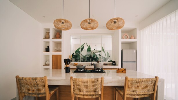 A modern kitchen with a central island, three wicker chairs, and a large window with plants. - Casa Nata Graceful 2 BR Mediteranian (Canggu)