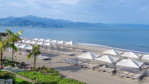 Ein Strand mit Reihen weißer Sonnenschirme und Liegestühle, freie Sicht auf das Meer und Berge im Hintergrund.