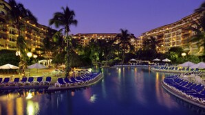 Ein Hotelpool mit Liegestühlen und Sonnenschirmen, umgeben von Palmen und Hotelgebäuden.