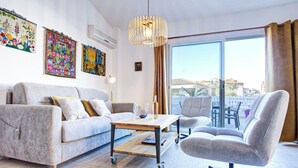 A modern living room with a sofa, a coffee table, and a chair. There are colorful wall hangings and a large window with a view. - Villa 12 Cassin by Interhome (Canet-Plage)
