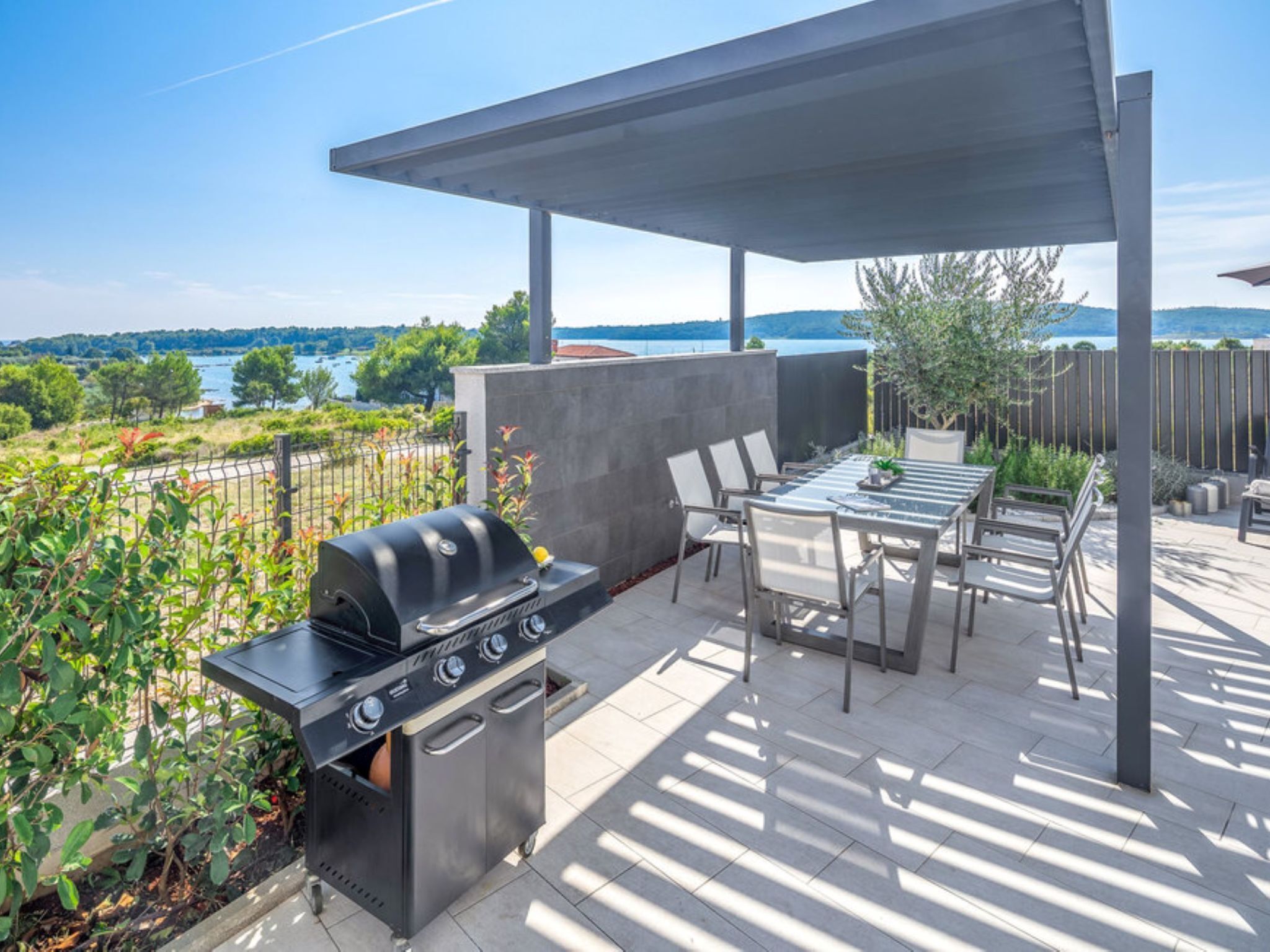 An outdoor patio with a grill, dining table, and chairs, overlooking a lake and hills.
