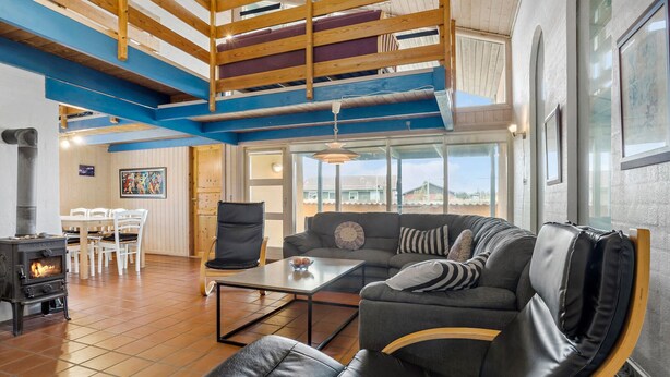 A spacious living room with a black leather sofa, a wooden coffee table, and a dining area with white chairs. A wood-burning stove is visible in the corner, and the room features exposed wooden beams and large windows. - Pet friendly home in Thisted with sauna (Thisted)