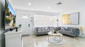A modern living room with a grey sectional sofa, a round coffee table, and a large window with blinds. - Stunning Home with a HEATED POOL 4 miles to the beach! (Pompano Beach)