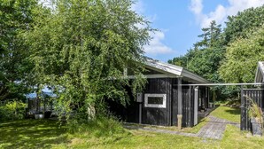 En lille, mørk træhytte med et enkelt vindue og en overdækket veranda, omgivet af frodig vegetation og træer.
