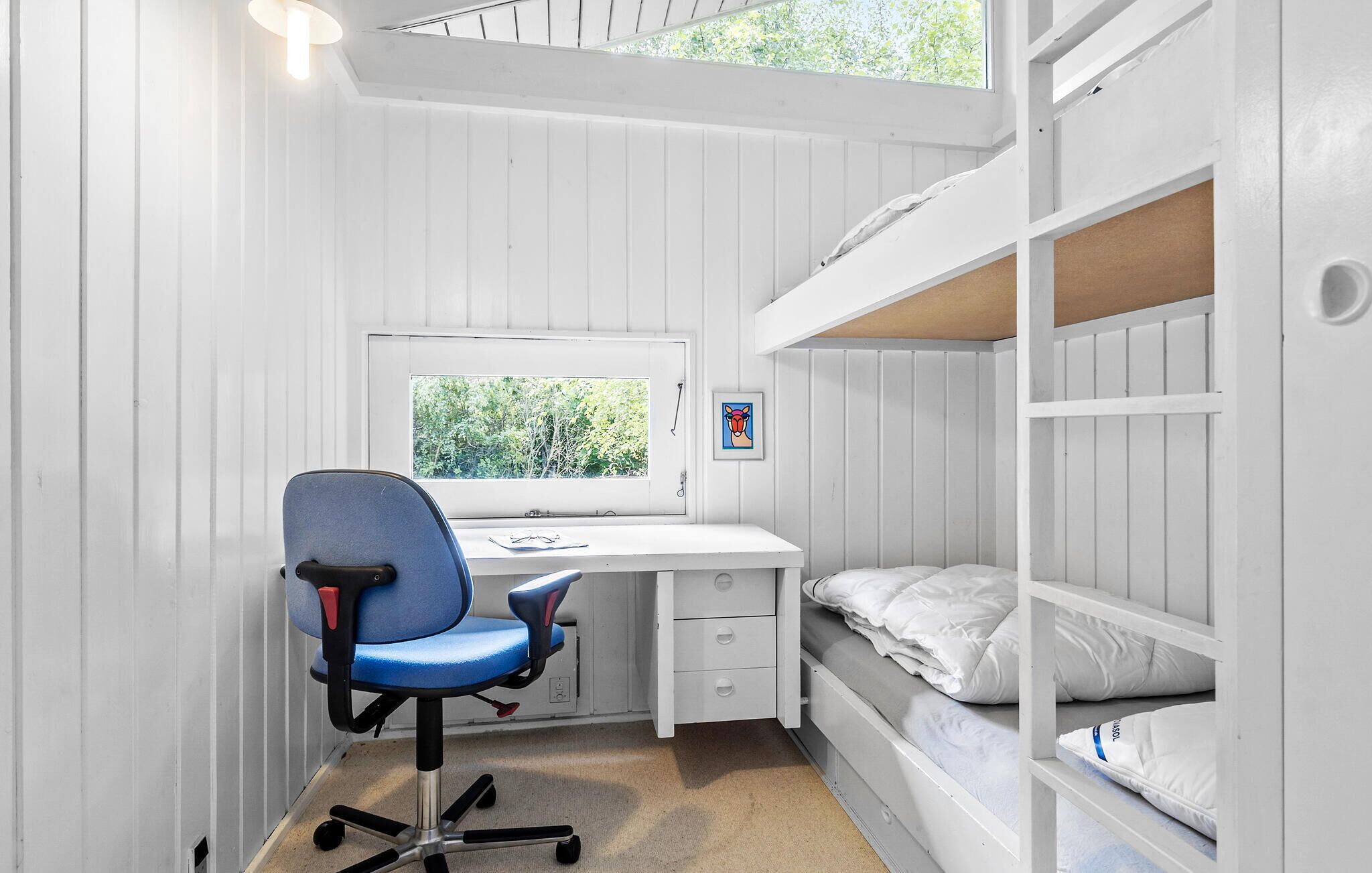 A compact room with a bunk bed, a desk with a chair, a window, and a framed picture on the wall.