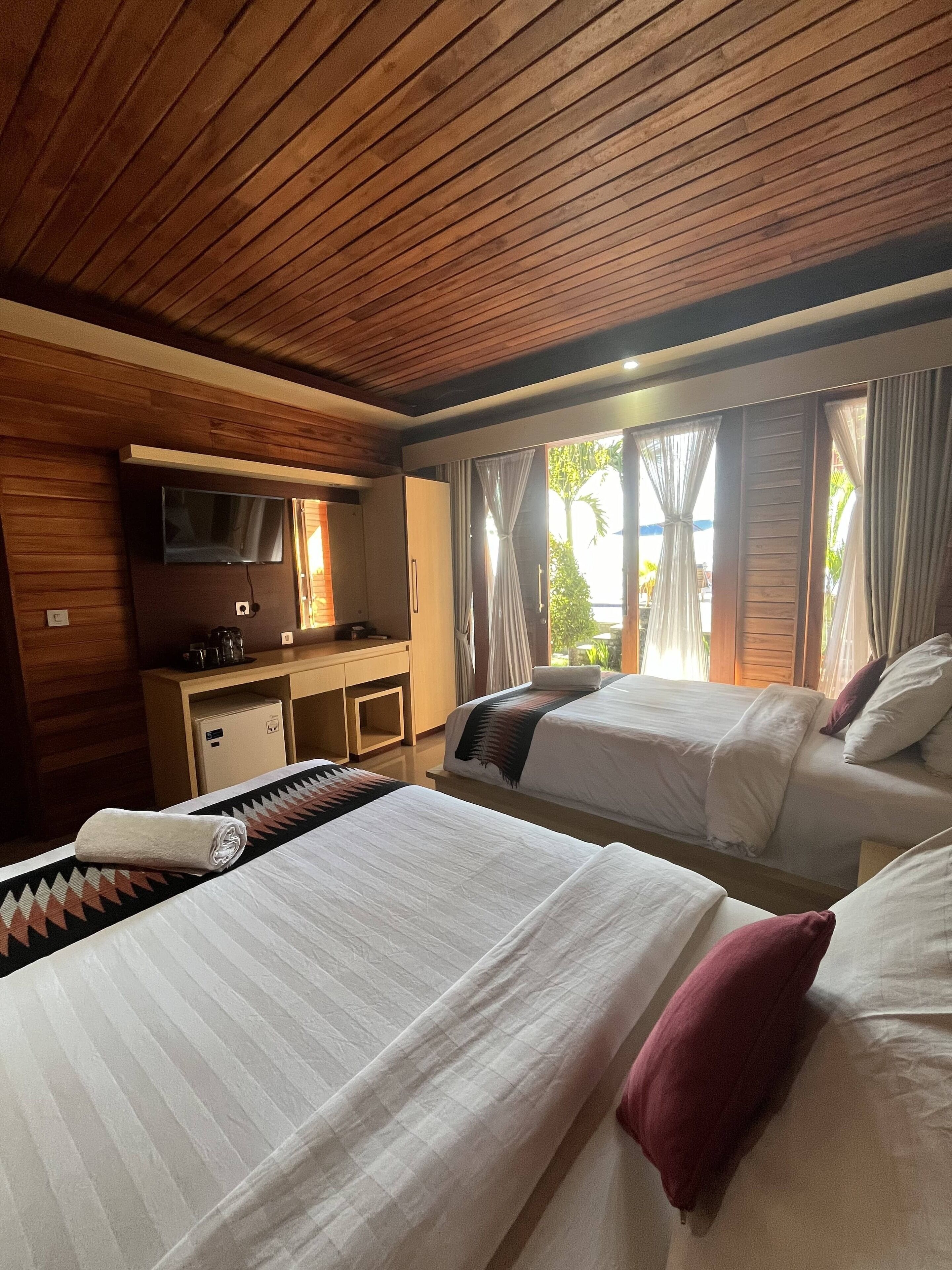 A hotel room with two beds, a TV, a small kitchenette, and large windows with curtains.
