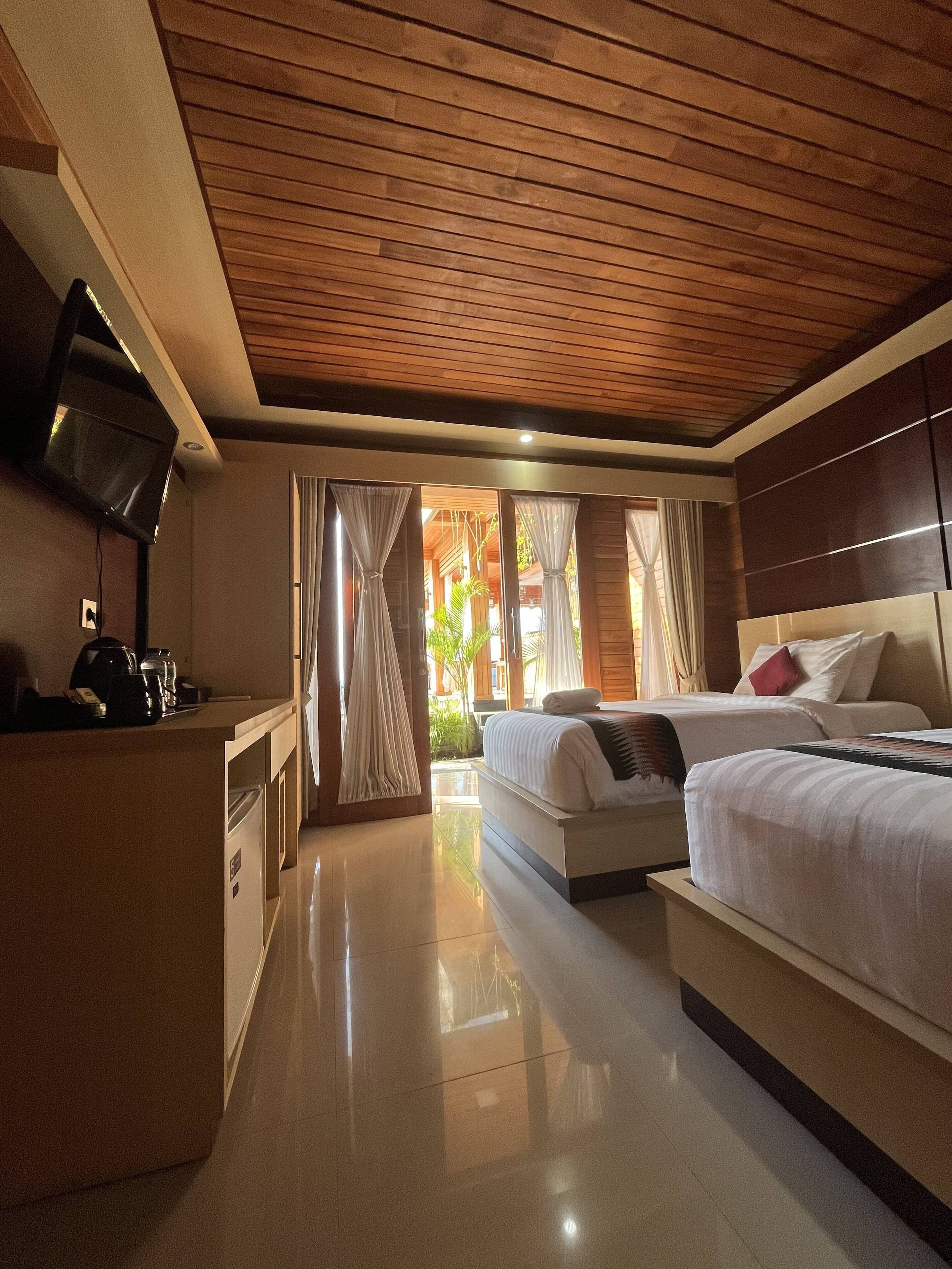 A hotel room with a wooden ceiling, a TV mounted on the wall, a bed with white bedding, and a small kitchenette area.