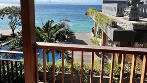 En træterrasse med udsigt over et klart, blåt hav, en traditionel bygning og en pool.