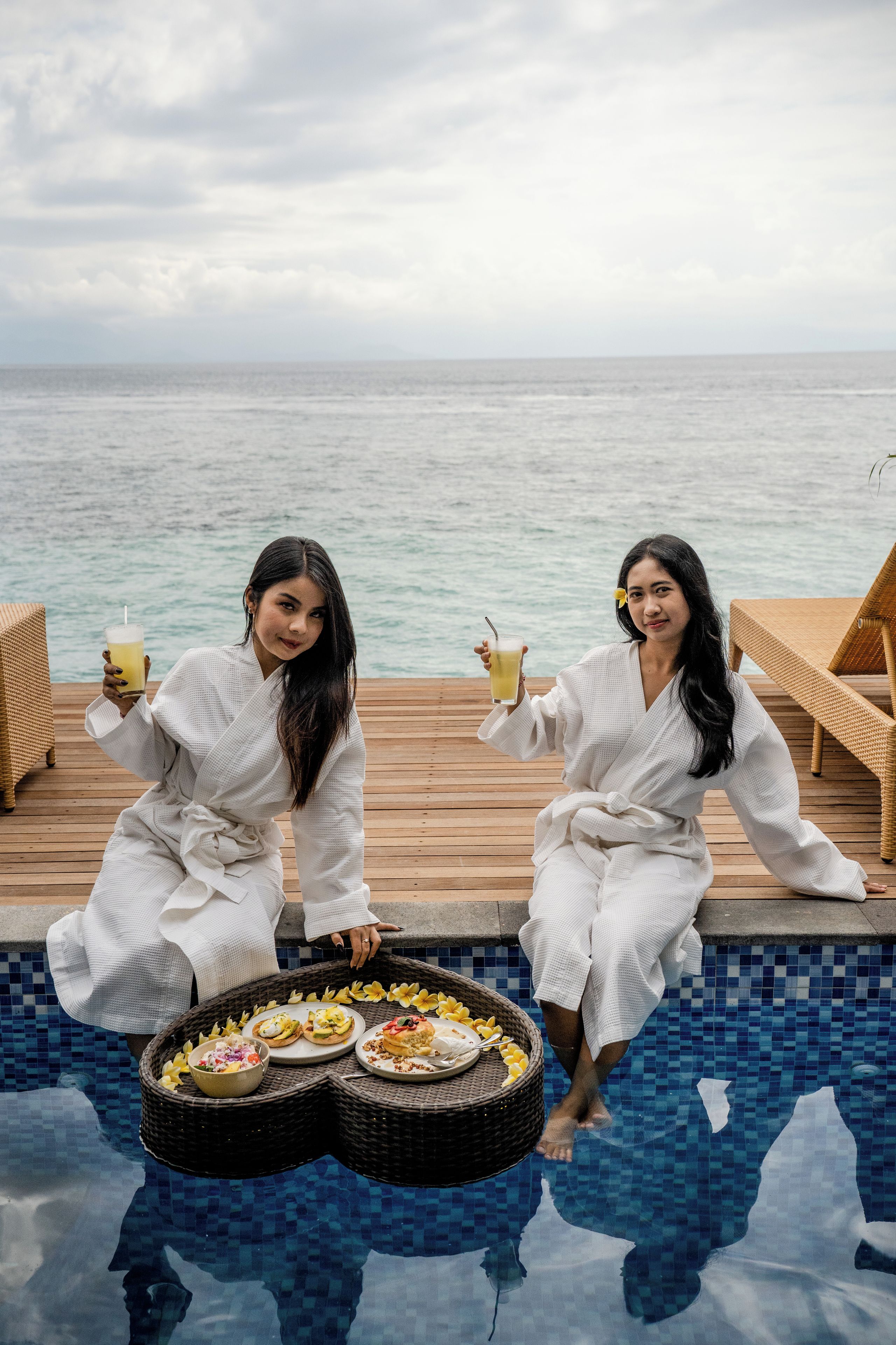 Duas pessoas sentadas à beira da piscina, com vista para o mar, segurando bebidas, e uma mesa com comida e uma cesta.