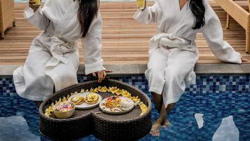 Two individuals seated by a poolside with a view of the sea, holding drinks, and a table with food and a basket.
