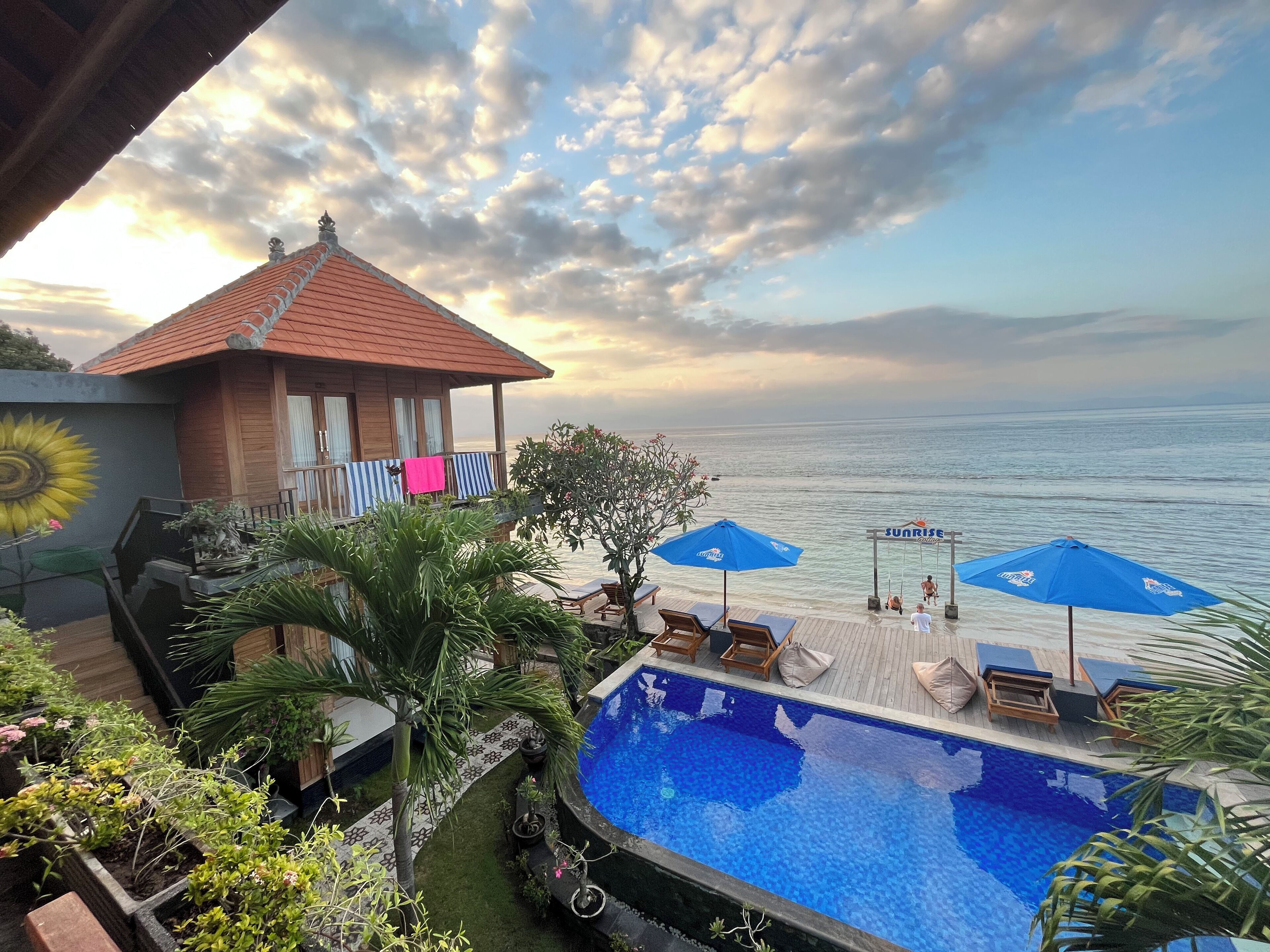 A pool area with a view of the sea, a wooden structure with a red roof, and a blue umbrella.