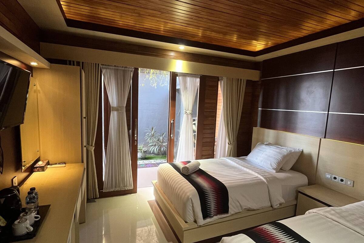 Une chambre d’hôtel avec un plafond en bois, un lit recouvert d’une courtepointe à motifs rouges et blancs, une table de chevet avec une lampe, et une vue sur l’extérieur grâce à de grandes fenêtres.