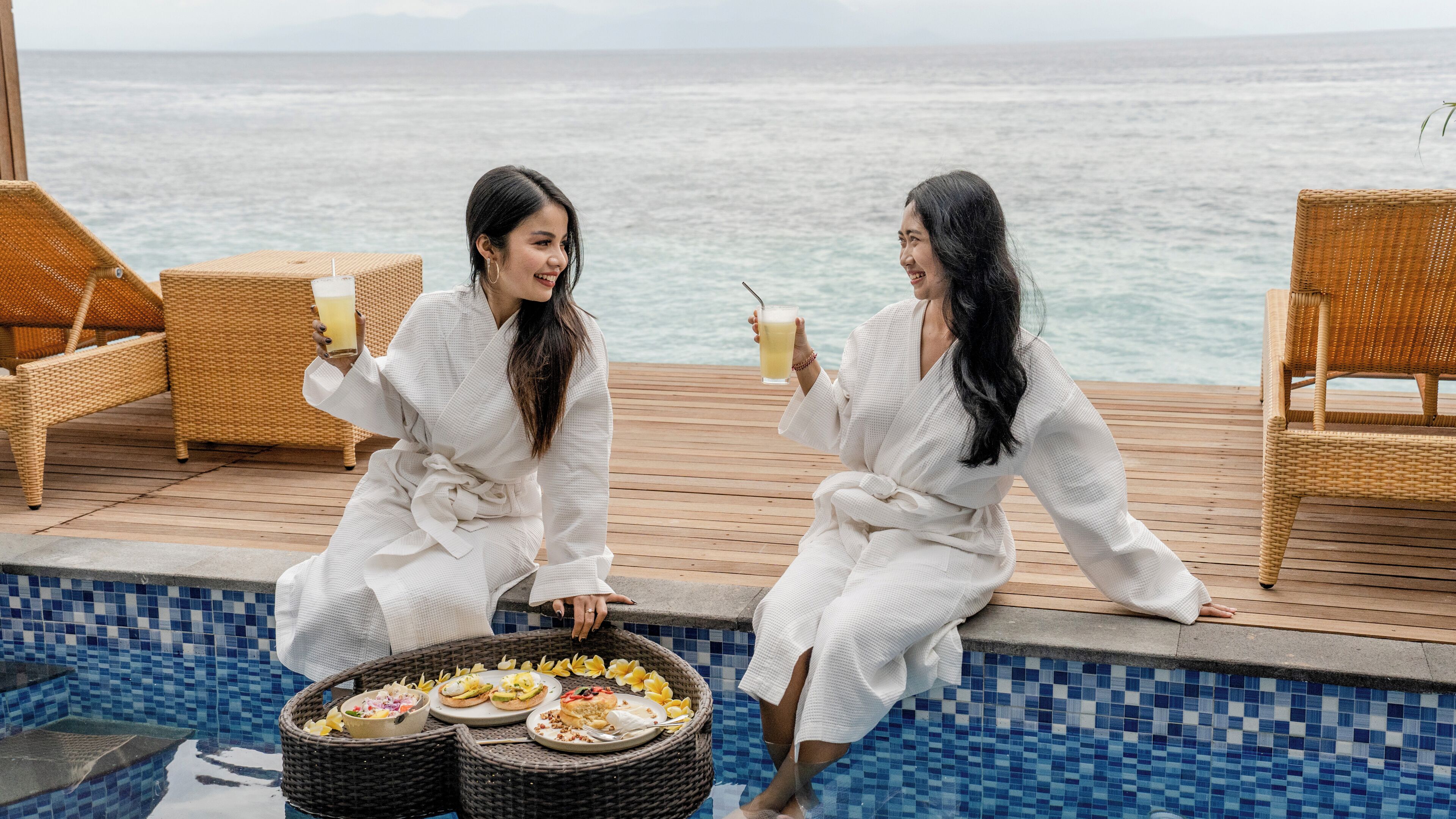 Dos personas vestidas de blanco sentadas junto a una piscina, con una silla de mimbre y una cesta de comida.