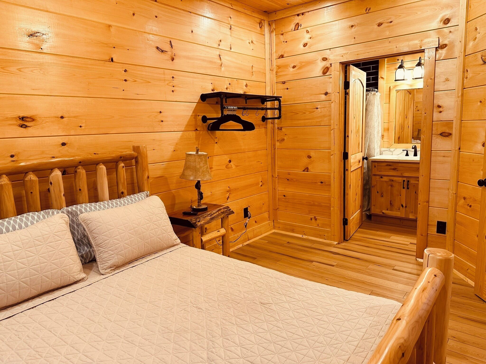 A wooden cabin bedroom with a bed, bedside table, lamp, and a hanging rack.