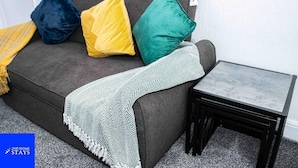 A grey sofa with a white and green herringbone throw blanket, a yellow pillow, a blue pillow, and a green pillow. A small black table with a grey top is beside the sofa. - Stoke House Sleeps 5 with Garden and Parking (Stoke-on-Trent)