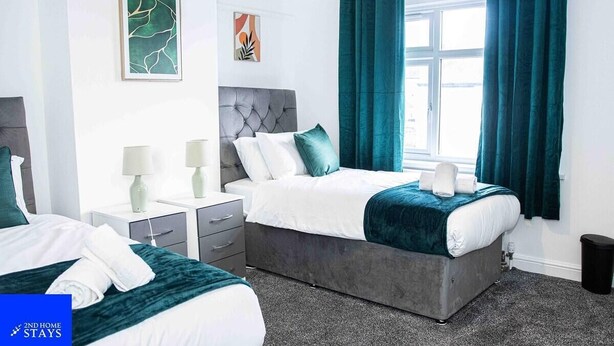 A hotel room with two beds, a grey upholstered headboard, teal curtains, and artwork on the walls. - Stoke House Sleeps 5 with Garden and Parking (Stoke-on-Trent)