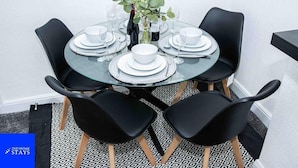 A round glass table set for four with black chairs, a vase with green plants, and a bottle of wine. - Stoke House Sleeps 5 with Garden and Parking (Stoke-on-Trent)