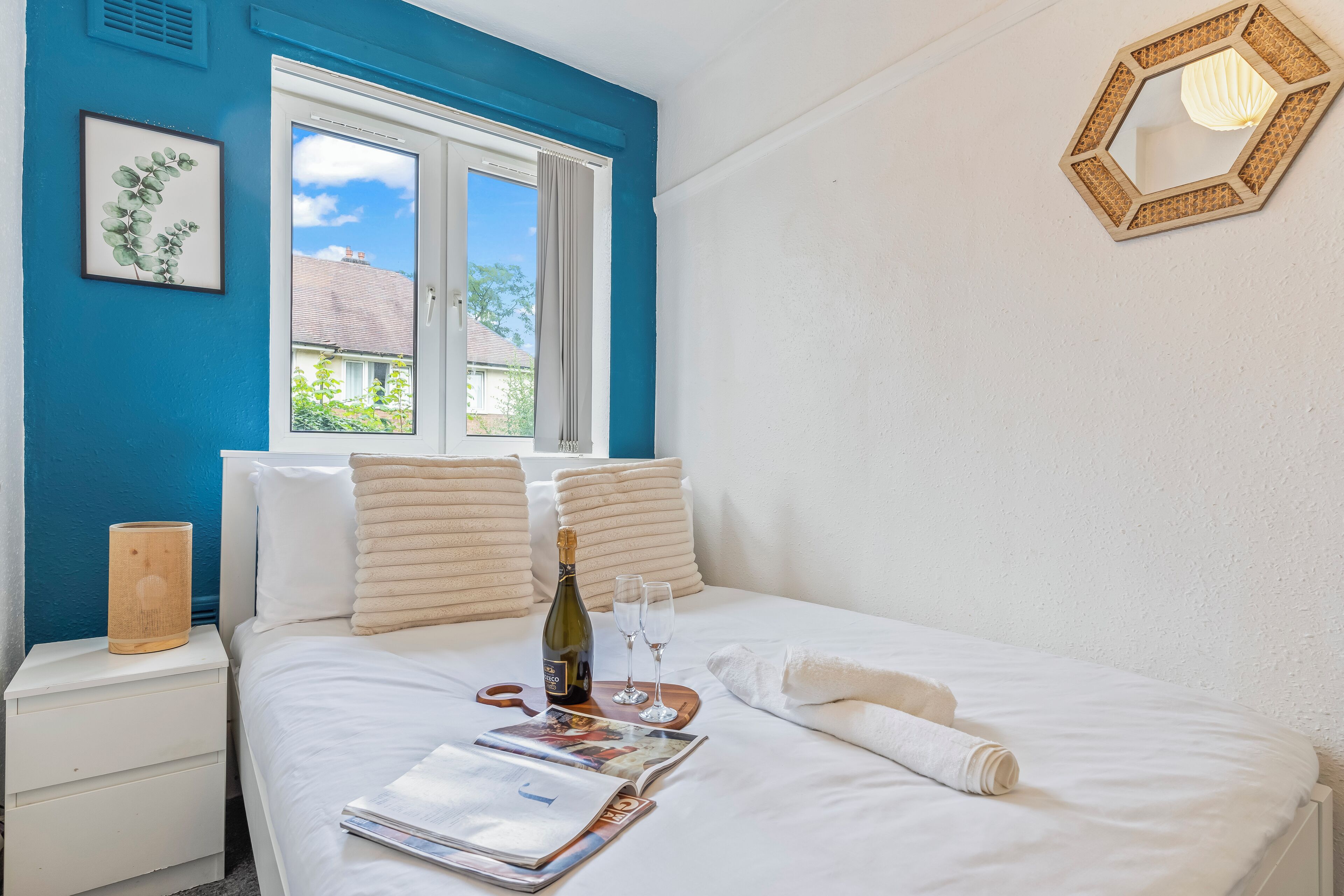 A neatly made bed with a bottle of champagne, two glasses, and a magazine on a bedside table.