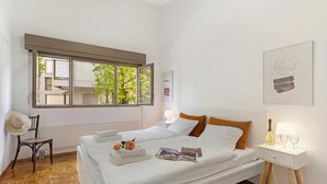 Ein Schlafzimmer mit großem Fenster, Nachttisch mit Lampe, einem Sessel und einem Bett mit Kissen und einer Zeitschrift.