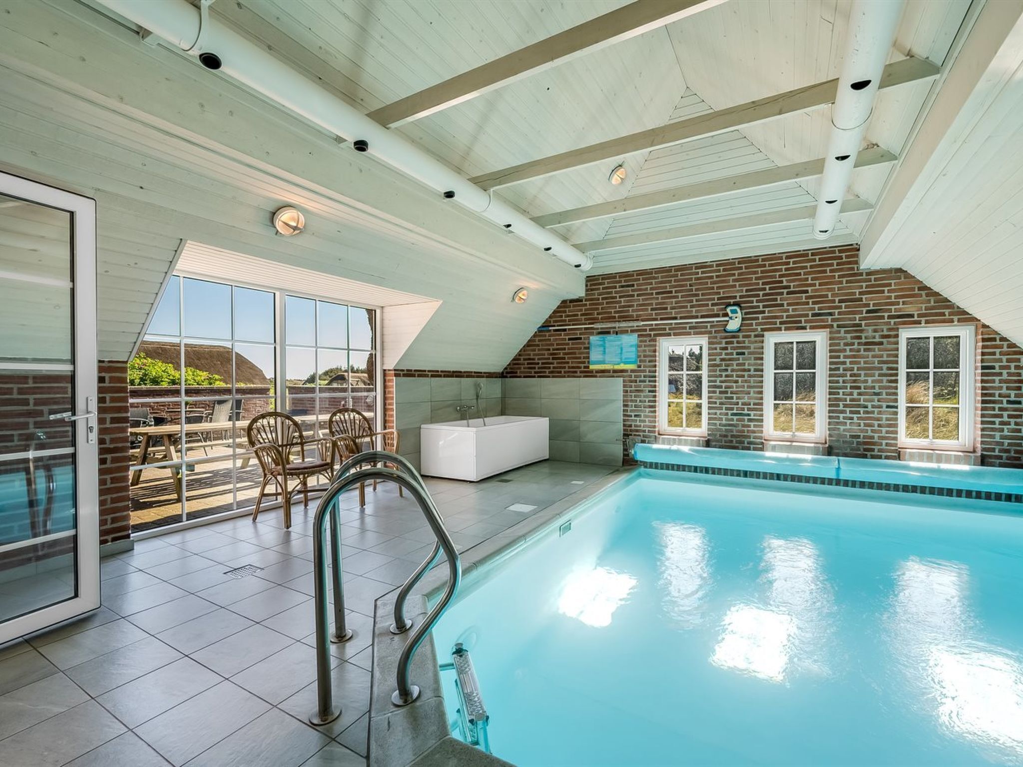 An indoor swimming pool with a clear view of the surrounding area, including a patio with outdoor seating and a view of the landscape.