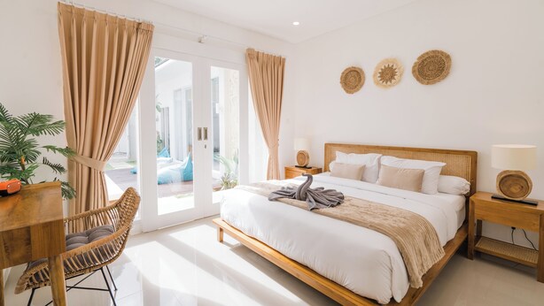 A bedroom with a wooden bed, a wooden nightstand, a wicker chair, and a dining table. - Villa Noni Kerobokan (Seminyak)