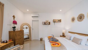 A hotel room with a bed, a wooden dresser, a mirror, a wall-mounted air conditioner, and a small shelf with decorative items. - Villa Noni Kerobokan (Seminyak)