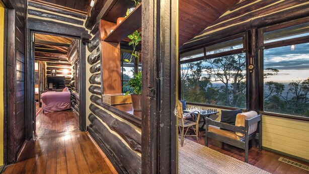 Interior de una cabaña de troncos con piso de madera, un sofá, una mesa de comedor y vista al exterior.