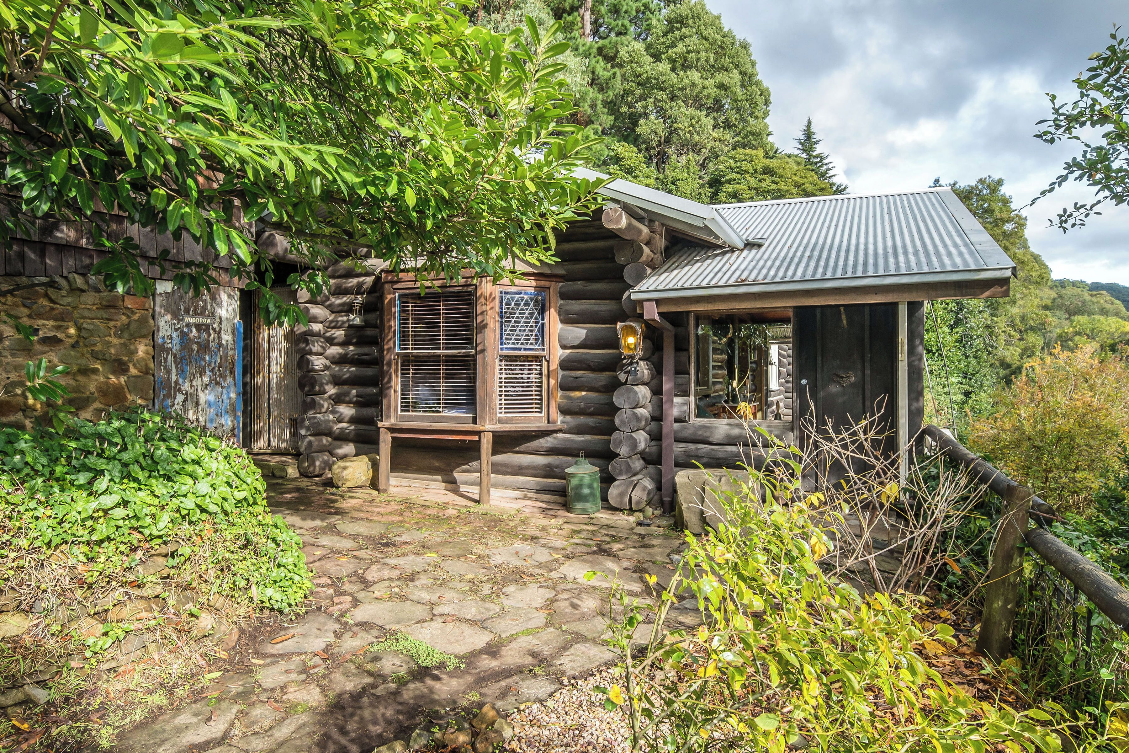 A rustic log cabin with a stone pathway leading to a wooden door, surrounded by lush greenery.