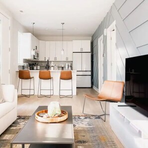 A modern kitchen with white cabinets, a dining area with three brown chairs, a sofa, and a flat-screen TV.