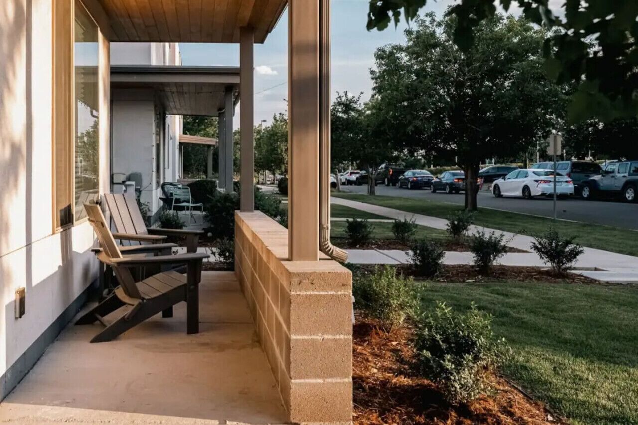 A covered outdoor seating area with chairs, a concrete block wall, and a view of a parking lot with cars and trees.