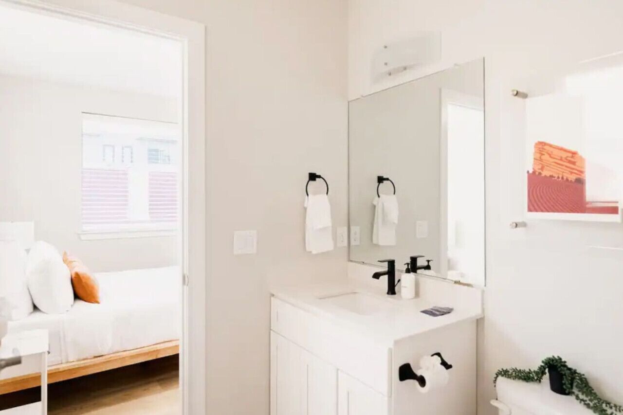 Une salle de bain moderne avec un meuble-lavabo blanc, un grand miroir et une fenêtre équipée de stores.